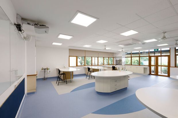 Bright, spacious commercial teaching or laboratory room with white walls, a suspended acoustic tile ceiling, recessed fluorescent lighting, extensive timber-framed windows and glazed doors, and multiple built-in workbenches and rounded central islands. The blue-and-off-white patterned resilient floor, sinks, wall-mounted projection equipment, and flexible stools suggest an educational science or technology classroom. The room has a clean, functional, contemporary institutional character.