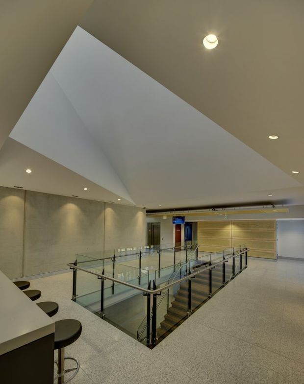 A modern commercial interior showing an open mezzanine or upper-level circulation area around a stairwell. The space features a large angular white ceiling plane, recessed downlights, pale concrete or plaster walls, terrazzo-like flooring, clear glass balustrades with dark metal posts, and timber-toned stair treads. A curved counter with black stools is visible at the left, while a service or lift zone appears at the rear.