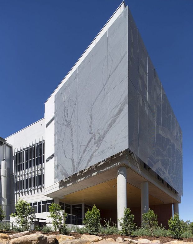 A contemporary multi-storey building exterior photographed from a low angle. The prominent projecting upper volume is clad in large pale grey panels with subtle veining, supported above a recessed ground-level entry or covered outdoor area by slender round columns. Adjacent white facade sections feature vertical metal sun-shading fins and horizontal window bands. Landscaping and exposed rocks sit at the base beneath a clear blue sky.