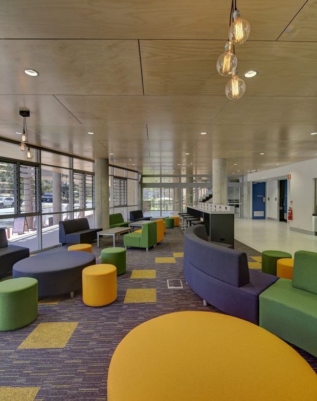A bright, contemporary commercial lounge or reception area with large louvred windows, a timber-panelled ceiling, polished concrete columns, and colourful modular seating. Curved purple and green sofas, yellow and green ottomans, and round tables sit on a patterned carpet with geometric yellow accents. Pendant globe lights and recessed downlights illuminate the open space, while a reception or service counter and blue doors are visible in the background.
