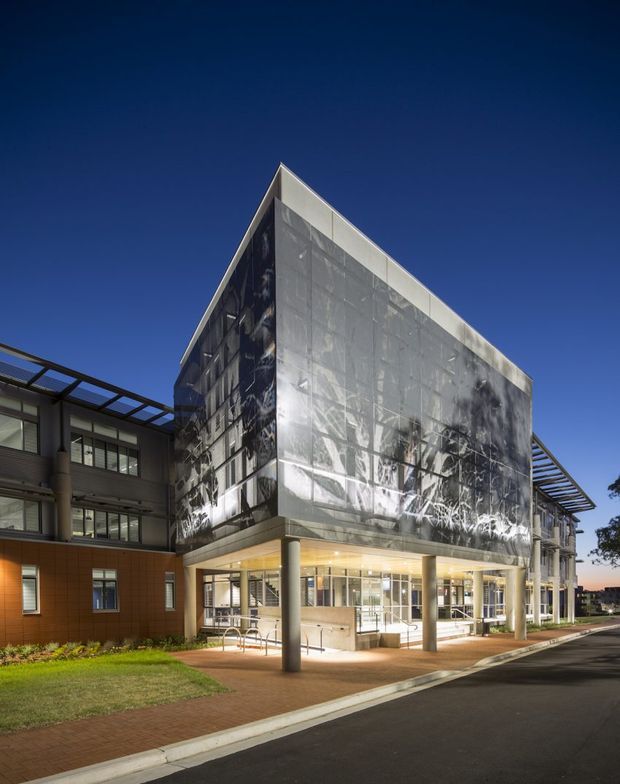 Exterior architectural photograph of a contemporary multi-storey institutional or commercial building at dusk. The main volume has a sharply angled roofline and a large translucent or reflective patterned facade, with glazed ground-floor entry areas, exposed structural columns, and projecting metal sun-shading canopies. Adjacent brick-clad wings and landscaped edges frame the illuminated entrance.
