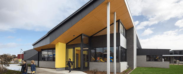 Exterior view of a contemporary institutional or commercial building with a strong projecting entrance canopy, warm timber soffit, dark metal roof and cladding, pale masonry walls, extensive glazing, and a bright yellow feature wall at the entry. The low-slung building sits beside landscaped lawn and paved paths under a partly cloudy sky.