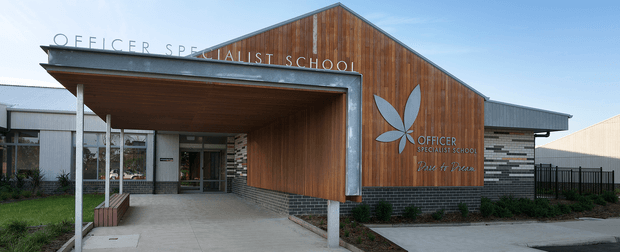 Front exterior view of a contemporary specialist school building. The entrance is sheltered by a broad flat canopy supported on slender posts, with a glazed doorway and office windows beneath. The main gabled facade combines warm vertical timber cladding with dark horizontal brick masonry, grey metal framing and roofing, and integrated school signage and logo. A paved approach, landscaped planting, fencing, and adjacent building wings are visible.