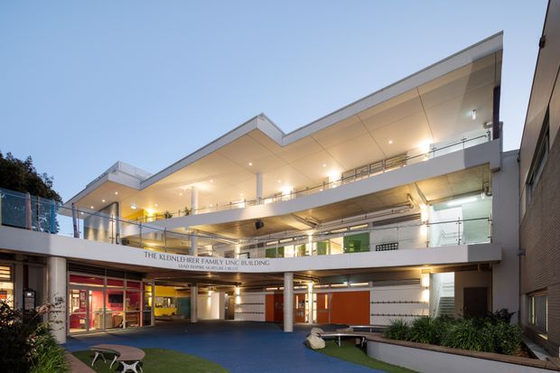 Exterior view of a contemporary multi-level institutional building at dusk, with open terraces, broad cantilevered slab edges, glazed circulation areas, integrated downlighting, and a covered ground-level courtyard. The facade combines white rendered surfaces, exposed concrete, glass balustrades, and colourful internal wall panels. Signage identifies the Kleinheinz Family Living Building.