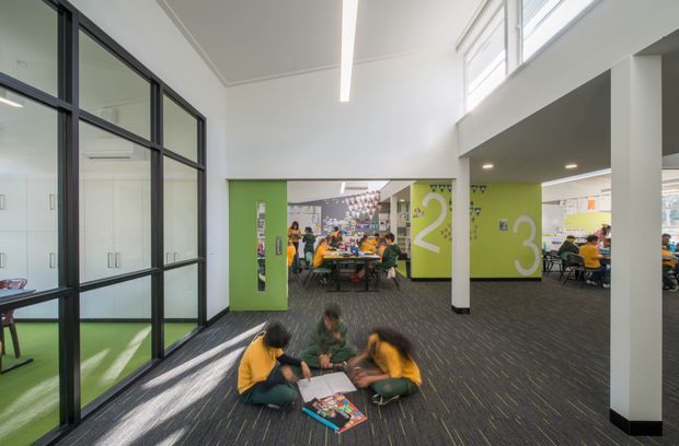 A bright, open-plan educational interior with children working on the floor in the foreground and collaborative tables in the background. The space features lime-green graphic walls marked with large white numbers, black-framed glazed partitions enclosing adjoining rooms, white structural columns, high clerestory-style glazing, linear LED lighting, and dark patterned carpet flooring.