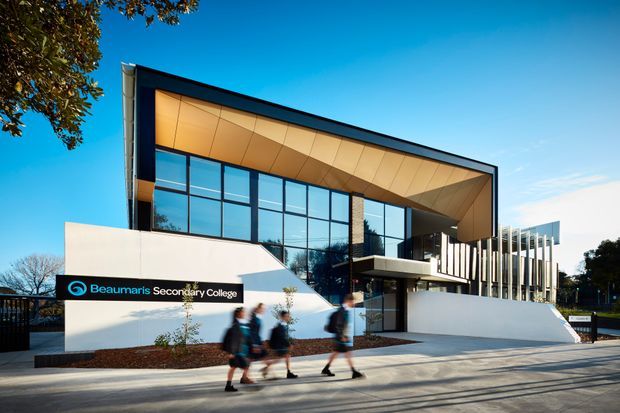 Exterior architectural photograph of Beaumaris Secondary College, showing a contemporary school entrance with a sharply angled roof canopy, expansive blue-tinted glazing, white rendered walls, dark framing, and vertical fins at the entry. Students walk along the paved frontage, with landscaped planting and trees surrounding the building.