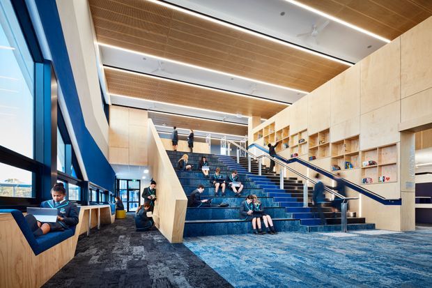 A large contemporary educational or institutional interior with a dramatic blue stair and tiered seating area, integrated timber-lined walls and extensive built-in shelving. Floor-to-ceiling windows line the left side, while linear recessed lighting and warm timber ceiling panels create a bright, open multi-level gathering and study space. Students are seated on the steps and at built-in window benches.