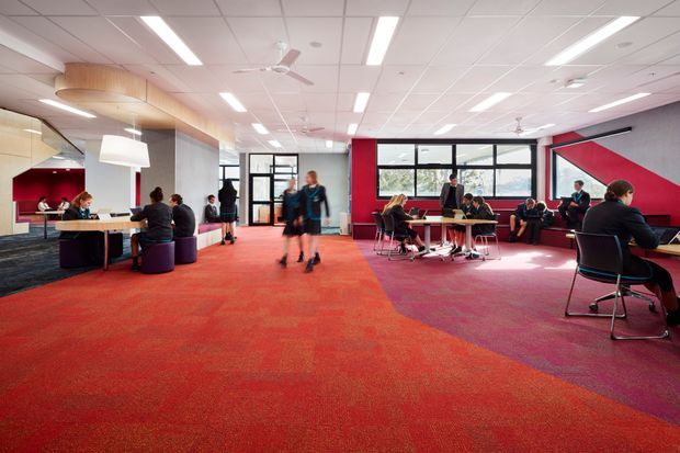 A bright, open-plan commercial lounge or collaborative workspace with vivid red carpet, scattered tables and chairs, booth seating, and people working or socialising. The room has extensive black-framed glazing, a suspended white tiled ceiling with linear fluorescent lighting and ceiling fans, plus bold red and dark grey geometric wall panels. A small kitchenette or service counter is visible at the rear left.