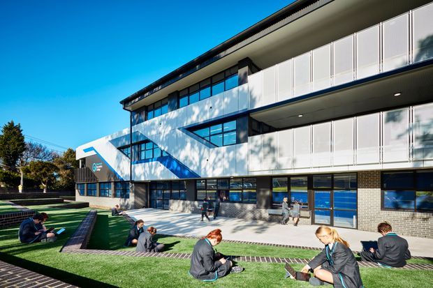 Exterior view of a contemporary multi-storey institutional or commercial building with extensive glazing, white vertical-panel balcony screens, dark brick lower walls, and a prominent angular white entrance canopy. Students are seated on tiered landscaped outdoor terraces in the foreground, with a clear blue sky and mature trees beyond.