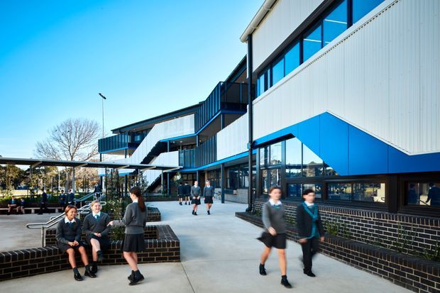 Exterior view of a contemporary multi-storey educational or commercial building with white and blue metal-clad facades, extensive dark-framed glazing, projecting balconies and angular volumes. Students and seating occupy a broad paved courtyard or forecourt, with covered outdoor structures and landscaped areas visible at the side.