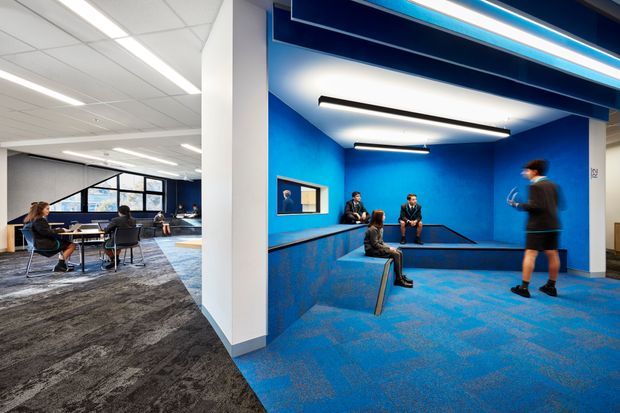 A contemporary commercial interior featuring an open meeting and collaboration area on the left and a vivid blue breakout or lounge zone on the right. The space has exposed grid ceilings, linear recessed and suspended lighting, blue-painted walls, blue patterned carpet, dark carpet in the meeting area, large interior windows, and built-in bench seating. Several people are using the workspace, with blue joinery and architectural framing creating a bold, energetic office environment.