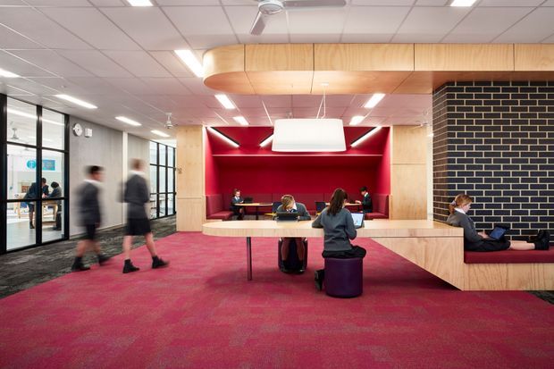 A contemporary commercial interior, likely a school or workplace commons, featuring an open collaborative seating and study area. The space has a vivid red carpet, pale timber-clad built-in bench seating and ceiling bulkhead, a dramatic red recessed alcove with integrated seating, a large suspended rectangular light, and a black brick feature wall. Glazed partitions and multiple people indicate an active shared workspace or campus lounge.