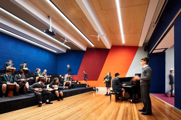 A contemporary music or performance rehearsal room with a seated group of students playing guitars and percussion on tiered black platforms, while a pianist performs at a grand piano. The room features bold blue, red, orange, and white geometric wall panels, a warm timber-panel ceiling with integrated linear lighting, ceiling fans, a polished timber floor, and large dark-framed glazing or doors along one side.