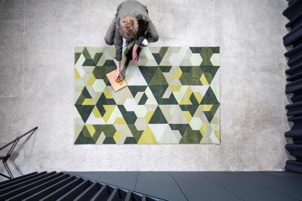 Overhead view of a modern interior showroom or gallery with a large geometric area rug in olive green, yellow, grey, cream, and white arranged on a pale concrete floor. A person is kneeling beside the rug, while dark architectural stair or railing elements frame the lower and right edges of the image.