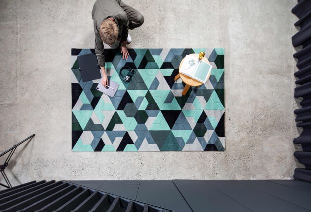 Overhead view of a contemporary interior workspace or lounge area with a geometric teal, charcoal, grey, and cream rug on a polished concrete floor. A person is seated or working at the rug beside a small round table with books and a decorative object. Dark sculptural stair or architectural elements frame the lower and right edges of the image.
