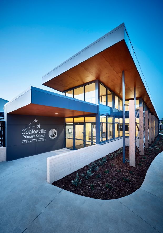 Exterior architectural photograph of Coatesville Primary School, showing a contemporary two-storey entrance building with strong horizontal roof overhangs, blue and white facade panels, extensive glazing, warm timber soffits, exposed slender steel columns, and a landscaped entrance forecourt with curved concrete paving and low white brick walls. School signage is visible on the dark entrance wall.