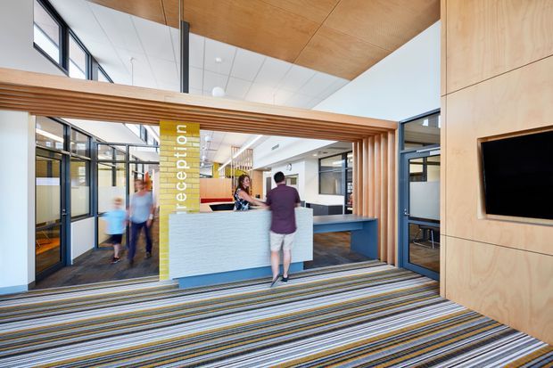 A bright contemporary commercial reception area with a central light-coloured reception desk, timber slatted portal framing the entry, pale timber wall panelling, extensive internal glazing, and a wall-mounted black display screen. The space has a high white ceiling with a warm timber ceiling panel and a striped commercial carpet floor. A vertical yellow reception sign and several people are visible.