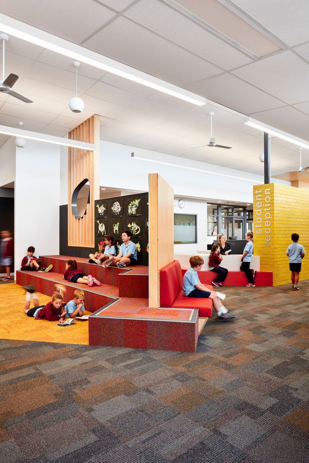 Bright contemporary school reception and student commons area with colourful tiered built-in seating, red upholstered benches, timber privacy screens, a yellow brick reception wall, patterned carpet flooring, suspended linear lights, and ceiling fans. Children and teenagers are using the informal seating and study areas.