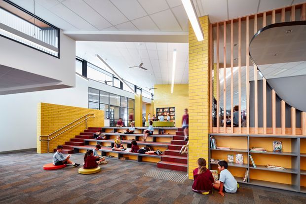 Bright contemporary school or educational library interior with stepped red-seated tiered platforms, children reading and working, yellow brick feature walls, extensive internal glazing, timber vertical screening, open shelving, suspended linear lighting, and a patterned carpet floor. The space has a spacious, airy, collaborative learning-centre character.