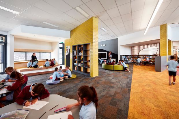 A bright, open-plan educational interior with students studying and socialising across multiple zones. The space features yellow brick piers with integrated shelving, large windows, built-in tiered seating, colourful curved lounge seating, timber slatted screening, carpet tile flooring, and a suspended grid ceiling with linear lighting.
