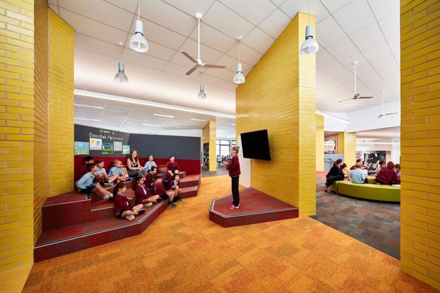 A bright institutional common area, likely a school or library, with yellow brick columns, tiered red seating, a central presentation platform with wall-mounted screen, groups of people, ceiling fans, and a patterned carpeted floor. The space has an open-plan collaborative layout and strong red, yellow, and orange colour accents.