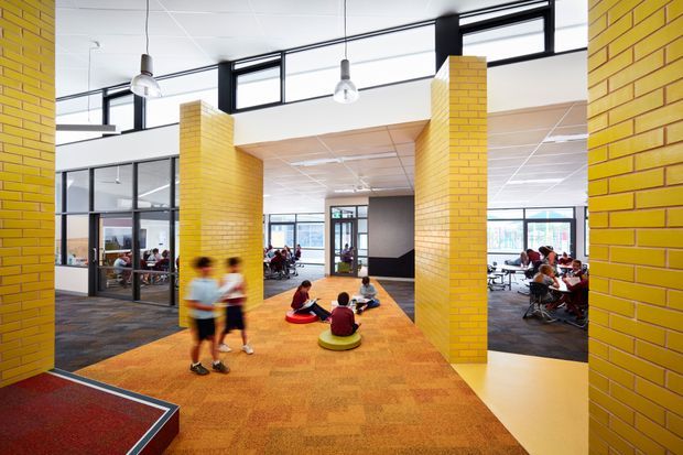 A bright, open-plan educational or community interior with vivid yellow brick columns framing a central carpeted circulation and gathering area. Children sit on colourful circular floor cushions, with additional seating and activity spaces visible through black-framed glazed partitions. High clerestory windows, suspended pendant lights, exposed ceiling grid, and patterned carpet create a contemporary institutional setting.