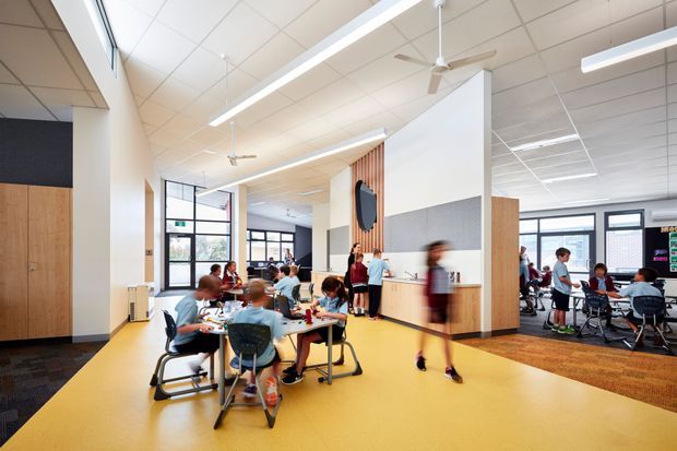 Bright, open-plan educational interior with students and tables arranged across a large shared learning space. The room features warm timber built-in cabinetry and vertical timber slats, pale acoustic wall panels, a yellow resilient floor, exposed suspended ceiling tiles, linear LED lighting, ceiling fans, and extensive dark-framed glazing. A partially enclosed classroom or breakout area is visible at the right.