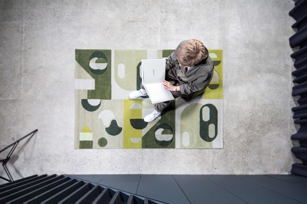 Overhead view of a person reading or working on a laptop while seated on a geometric patterned rug in a concrete-floored interior. Dark metal stair or architectural elements frame the lower and right edges of the composition.
