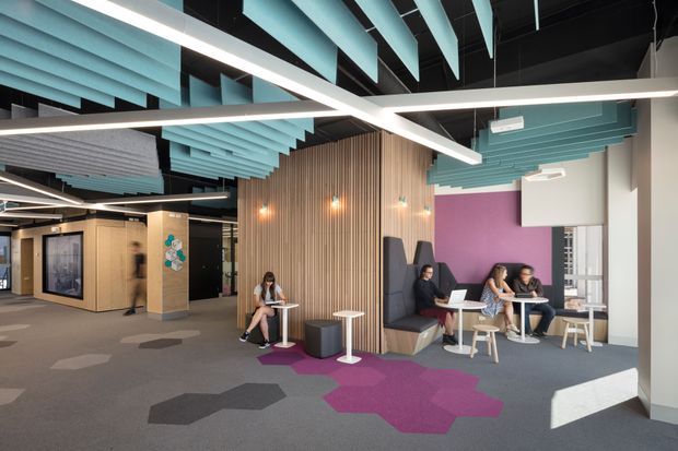 A contemporary commercial lounge and breakout workspace with informal seating, small round tables, booth seating, and people working on laptops. The open-plan interior features a dark exposed ceiling, suspended linear lighting, turquoise acoustic ceiling baffles, vertical timber slat wall cladding, a vivid purple accent wall, and patterned grey and magenta carpet tiles.