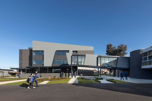 Wide exterior view of a contemporary multi-storey institutional or commercial building complex. The architecture features rectilinear volumes, large glazed curtain walls, pale panel cladding, dark metal facades, projecting canopies and landscaped pedestrian approaches. People are visible near the entrance, with a clear blue sky and paved forecourt.