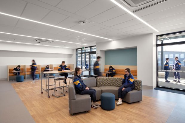 A bright contemporary student common area or lounge with light timber-look flooring, modular grey upholstered seating, blue cylindrical ottomans, study desks, booth seating, and students using the space. The room has white acoustic ceiling panels with linear LED lighting, large glazed doors and windows, and a muted blue feature wall with built-in timber bench seating.
