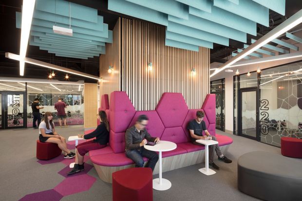A vibrant contemporary commercial interior, likely a coworking or office reception and lounge area. The space features clustered magenta upholstered seating with high geometric privacy backs, small white pedestal tables, red ottomans, grey carpet with magenta geometric inlays, and a dramatic suspended ceiling installation of layered aqua acoustic panels. Vertical timber slat wall cladding forms a warm feature backdrop, while glazed meeting rooms and glass partitions surround the seating area. Linear LED lighting, exposed dark ceiling elements, and bold graphic glazing add a modern workplace character.