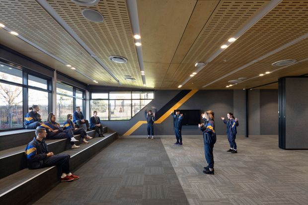 A contemporary school or institutional multipurpose interior with tiered built-in seating along the windowed wall and a group of students gathered in an open activity area. The room features dark grey walls with bold yellow and blue geometric graphics, expansive black-framed glazing, grey patterned carpet, and a suspended perforated timber-look ceiling with recessed lighting and circular HVAC diffusers.