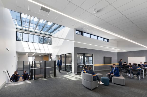Bright contemporary school interior with an open multi-level commons or student lounge. The space features a large skylight and clerestory glazing, white acoustic ceiling panels, dark carpet flooring, dark textured wall finishes with a horizontal yellow accent band, glass balustrades, and clusters of lounge seating and dining tables. Students and staff are present, with a stair or circulation zone visible to the left.