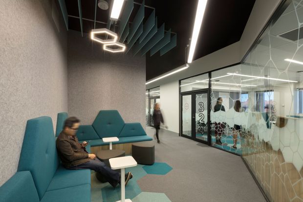 Contemporary commercial interior corridor with a flexible lounge and waiting area. Teal upholstered modular seating, small round and square tables, and geometric teal floor inlays sit against a textured grey wall. A black ceiling with linear LED lighting and suspended geometric acoustic fins creates a modern workplace atmosphere. Glazed offices and meeting rooms line the opposite side, with patterned privacy film on the glass and visible wayfinding graphics.