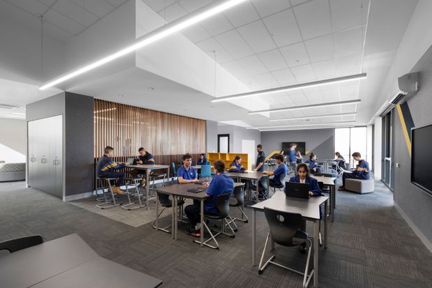 A bright contemporary commercial learning or collaboration space with students seated at shared tables using laptops and working together. The room has a high suspended acoustic-tile ceiling, linear pendant lighting, grey carpet flooring, white and charcoal walls, large windows, built-in bench seating, and a timber slat feature wall. A kitchenette or utility zone is partially visible at the rear.