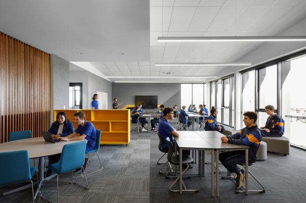 A bright, contemporary open-plan school or institutional learning commons with students working at tables and in small groups. The space features floor-to-ceiling glazing along one side, grey carpet tiles, white acoustic ceiling panels with linear suspended lighting, a large media screen, and vibrant blue and yellow built-in shelving and furniture. Vertical timber battens form a feature wall at the left.