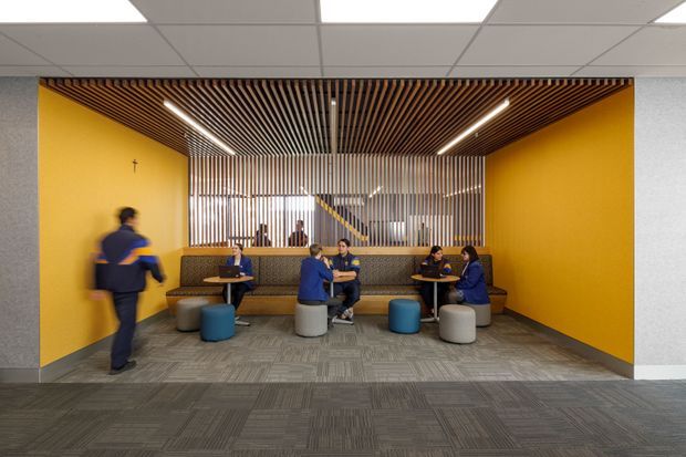 A bright contemporary commercial breakout lounge or staff collaboration area, framed by vivid mustard-yellow walls. The space contains built-in upholstered banquette seating, small round café tables, stools and poufs, with people using the area. A timber slatted ceiling feature and matching vertical screen create a warm architectural focal point, while linear strip lights illuminate the seating zone. The floor is covered in grey patterned carpet tiles and the surrounding ceiling uses a suspended acoustic grid.
