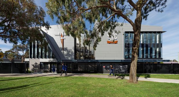 Exterior view of a contemporary institutional or commercial building set behind a broad lawn and mature trees. The facade combines pale grey panels, extensive vertical glazing, dark metal fencing and cladding, and a prominent angled structural element. A central entrance and signage are visible, with pedestrians on the footpath. The composition emphasizes the building frontage and landscaped setting.