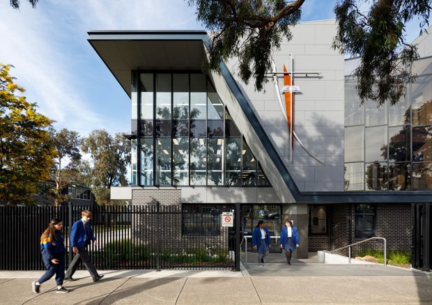 Exterior view of a contemporary multi-storey institutional or commercial building, featuring a sharply projecting flat roof, extensive floor-to-ceiling glazing, white panel cladding, dark brickwork, black metal fencing, and a prominent diagonal structural or facade element. People are visible near the entrance, with mature trees around the site.