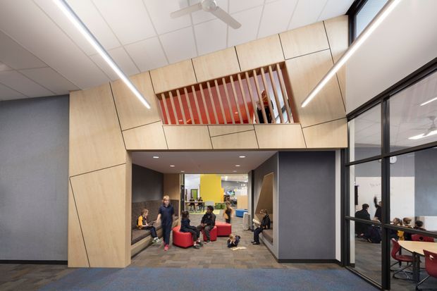 Interior view of a contemporary commercial communal area, likely an educational or workplace facility. A large light timber-clad portal frames a recessed lounge with red seating, while a slatted timber mezzanine balustrade and enclosed upper-level space sit above. Dark grey wall panels, exposed glass-partitioned rooms, linear LED lighting, carpet tiles, and a suspended ceiling create a modern institutional fit-out. People are present in the lounge and visible through the glazed meeting or classroom area.