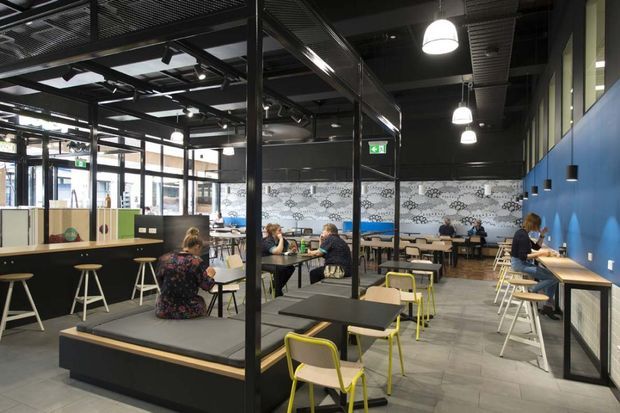 A spacious industrial-style café or hospitality interior with multiple communal and individual dining tables, black steel structural framing, exposed services and dark mesh ceiling, pendant lights, extensive glazing, a bold blue feature wall, patterned tiled wall panels, and light grey tiled flooring. The space is occupied by patrons and includes varied seating zones and built-in bench seating.