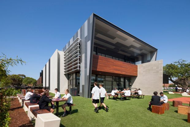 Contemporary multi-storey institutional or commercial building with a dramatic angular roof canopy, exposed structural geometry, vertical fins, extensive glazing, and a projecting timber-clad upper-level balcony. The building opens onto a landscaped courtyard or lawn populated with outdoor tables and seating, suggesting a campus, school, or hospitality setting.