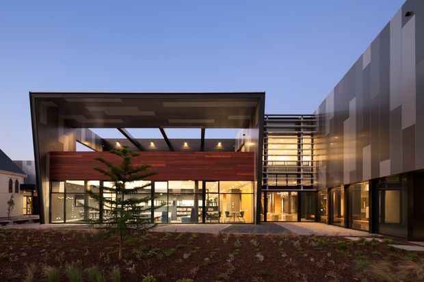 Twilight exterior view of a contemporary multi-storey building with a glazed ground floor, deep projecting rectangular canopy, warm timber-clad upper facade, extensive curtain-wall glazing, and a tall darker vertical-panel wing. A landscaped planted area and paved approach sit in the foreground.