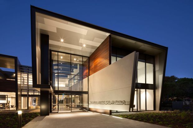 Twilight exterior view of a contemporary multi-level building with a dramatic angular entrance canopy and projecting wall forms. Extensive floor-to-ceiling glazing reveals illuminated interior spaces, while dark metal framing, warm timber wall cladding, and pale rendered or panelled surfaces create a refined modern facade. A paved approach and landscaped planting lead to the glazed entry.