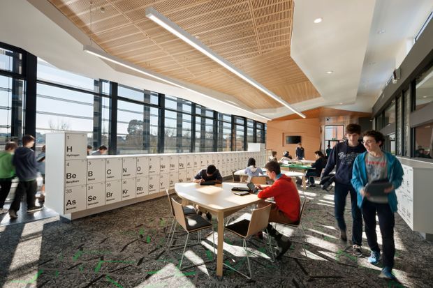 A bright, contemporary educational interior resembling a school or university learning commons. The large open room has floor-to-ceiling black-framed glazing, a dramatic slatted timber ceiling with linear lighting, white sculptural ceiling forms, collaborative tables and chairs, and a prominent periodic-table graphic partition. Students occupy the space, which has a lively, informal study atmosphere.