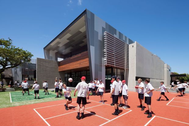 Exterior view of a contemporary multi-storey school or institutional building with a dramatic angular roofline, exposed upper-level terrace, concrete and metal facade elements, timber screening, and horizontal louvres. Students are playing on red and green outdoor courts in the foreground under bright daylight.