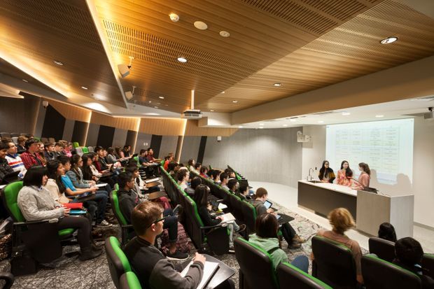 A modern tiered lecture theatre or seminar room viewed from the audience seating toward a presentation area. Rows of upholstered green and black seats face a long presentation desk with several speakers, a large projected screen, and a ceiling-mounted projector. The room features a warm slatted timber acoustic ceiling, recessed lighting, dark wall panels, grey acoustic wall surfaces, and patterned carpet. The space is occupied by an audience attending a presentation.