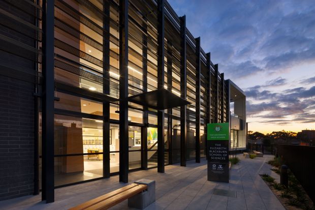 Exterior dusk view of a contemporary multi-storey institutional or commercial building. The facade features extensive floor-to-ceiling glazing, dark metal framing, projecting vertical fins, and warm timber-toned screening. A paved pedestrian forecourt with built-in bench seating leads to the illuminated entrance, while signage identifies the building as the Elizabeth Blackburn School of Medical Sciences. The surrounding urban setting and sunset sky are visible in the background.