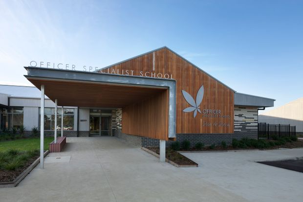 Exterior view of a contemporary specialist school entrance, featuring a prominent timber-clad gabled facade, broad covered entry canopy, exposed metal trim, glazed entry doors, and low stone-clad walls. The building signage identifies it as the Officer Specialist School.